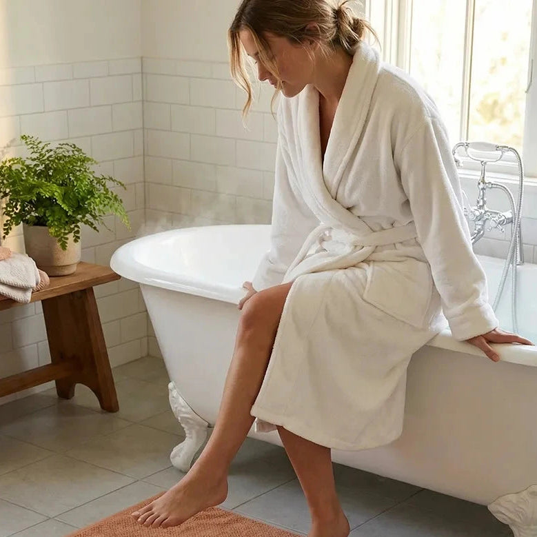 Woman in a white robe stepping onto a terracotta bath mat in a bathroom.