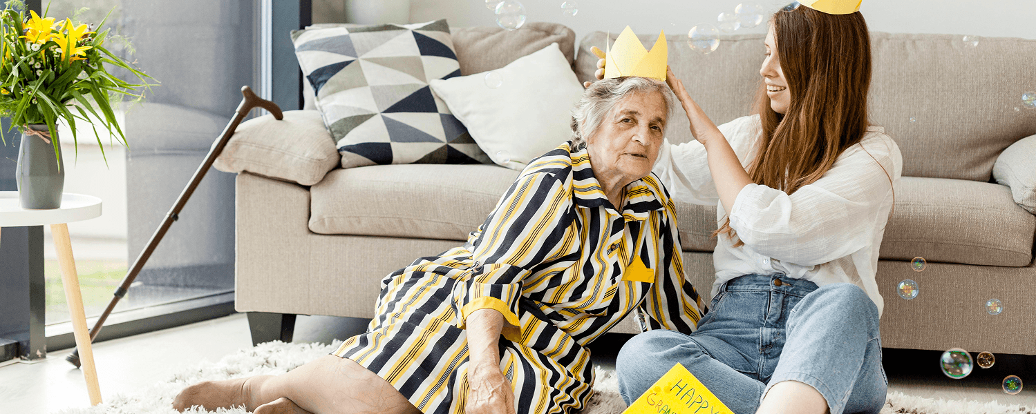 Smiling woman and elderly lady with paper crowns sitting on a plush white area rug in a cozy living room with bubbles floating and a bouquet of yellow flowers nearby.