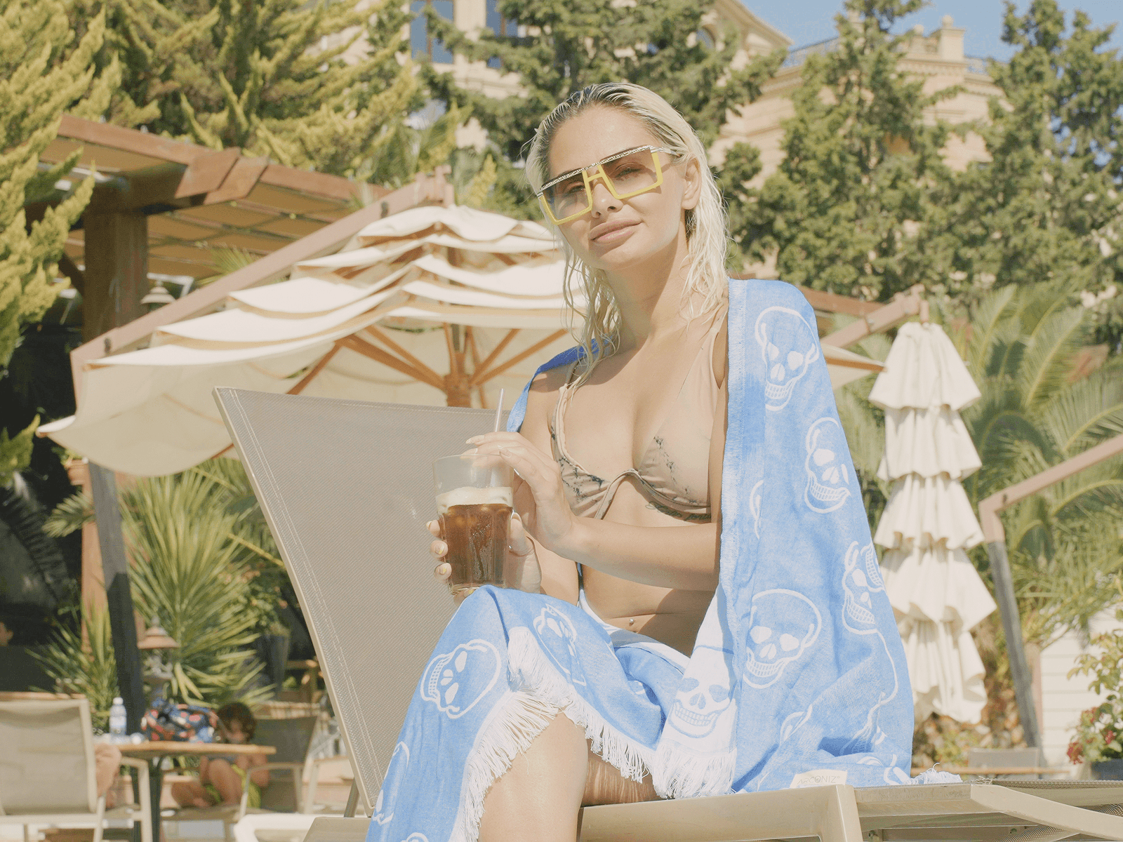Woman by pool lounging in bikini with blue beach towel and sunglasses, holding a cold drink
